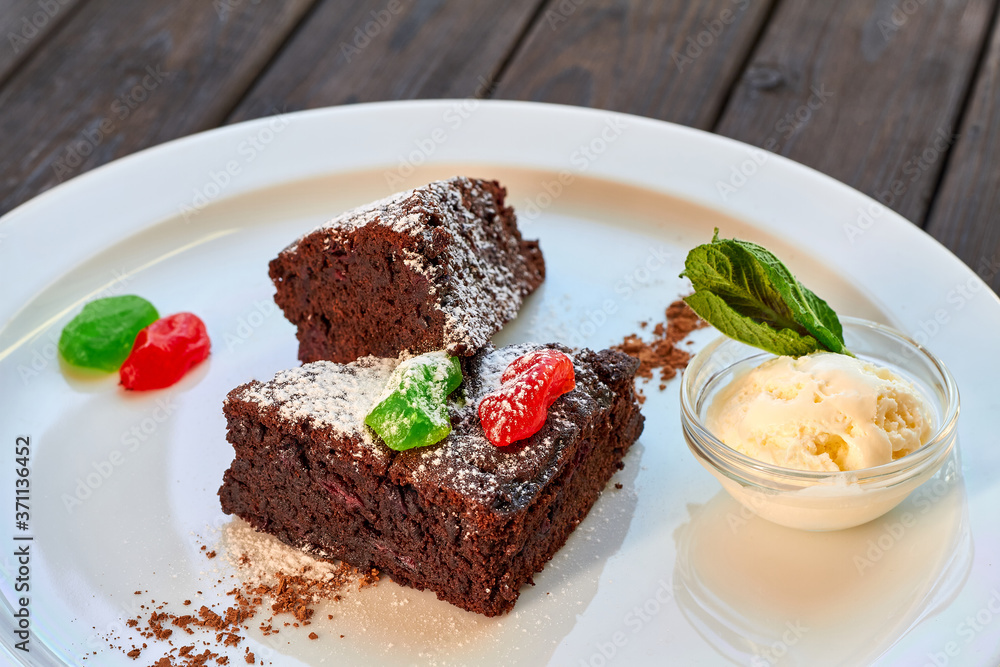 Slice of chocolate cake with candied fruit, mint and ice cream on a white plate and wooden background