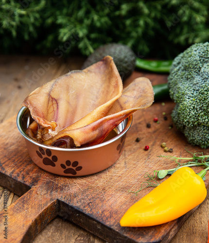 Dried pig ears in the dog vessel among the greenery on the wooden board. Chewing treats for domestic dogs. Natural dental treats for promotion and pampering.