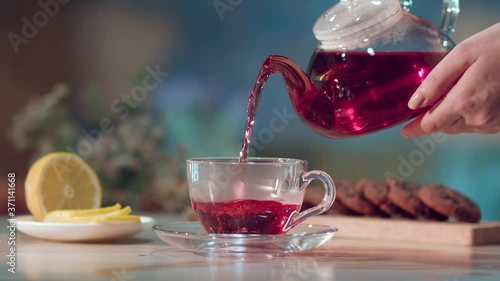 Woman's hand pouring tea from a transparent glass jug into a transparent cup of karkade tea with hibiscus flower. Breakfast concept. Lemon slices. Tea time.