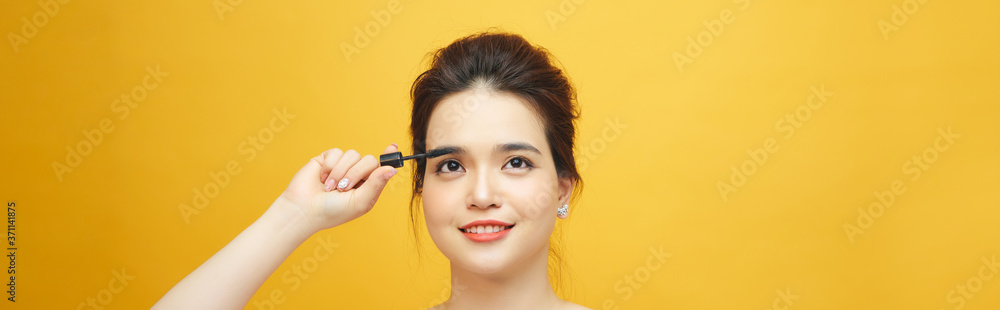 Closeup portrait of attractive young woman putting some mascara onto her eyelashes with make up brush over yellow background