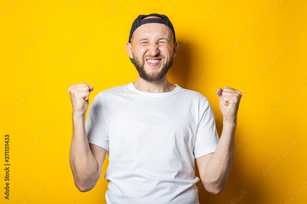 Bearded young man in baseball cap with closed eyes rejoices holding his fists on yellow background. Bearded young man in baseball cap with closed eyes rejoices holding his fists on yellow background.