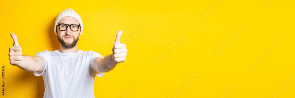 Smiling affable young guy with a beard in a cap and glasses shows a gesture with his hands, thumbs up, class on a yellow background. Banner. Smiling affable young guy with a beard in a cap and glasses shows a gesture with his hands, thumbs up, class on a yellow background. Banner.