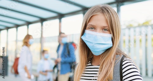 Portrait of pretty Caucasian blonde cute teen girl in medical mask standing at bus stop outdoor and looking at camera. Zooming in. Parents with small brother and suitcases on blurred background.