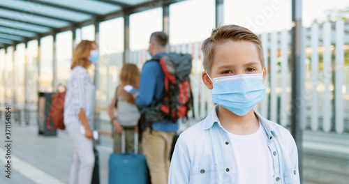 Portrait of Caucasian cute teen boy in medical mask standing at bus stop outdoor and looking at camera. Zooming in. Parents with pretty sister and daughter and suitcases on blurred background.