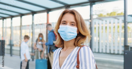 Portrait of beautiful Caucasian blonde woman in medical mask looking at camera and standing at bus stop or train station. Husband with kids and suitcases waiting for transport on blurred background.