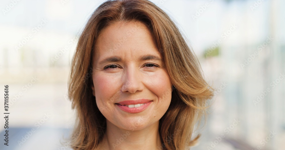 Portrait of beautiful Caucasian woman with fair hair looking at side, turning face to camera and smiling outdoors on sunny summer day. Happy female at street. Close up. Portrait of beautiful Caucasian woman with fair hair looking at side, turning face to camera and smiling outdoors on sunny summer day. Happy female at street. Close up.