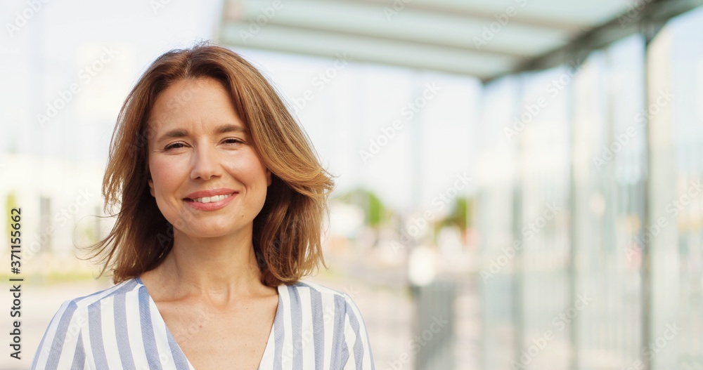 Portrait shot of beautiful Caucasian woman with fair hair looking at side, turning face to camera and smiling outdoors on sunny summer day. Happy female at street. Close up of cheerful lady. Portrait shot of beautiful Caucasian woman with fair hair looking at side, turning face to camera and smiling outdoors on sunny summer day. Happy female at street. Close up of cheerful lady.