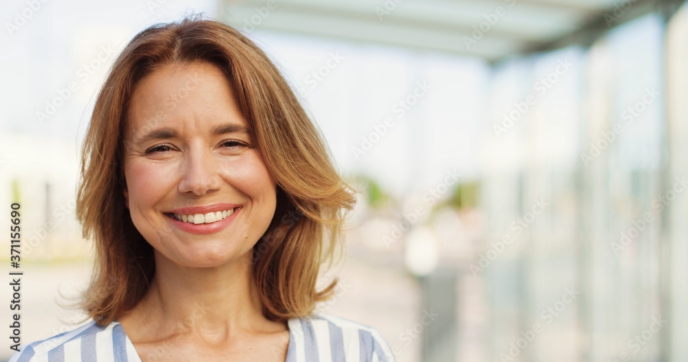 Portrait shot of beautiful Caucasian woman with fair hair looking at side, turning face to camera and smiling outdoors on sunny summer day. Happy female at street. Close up of cheerful lady.
