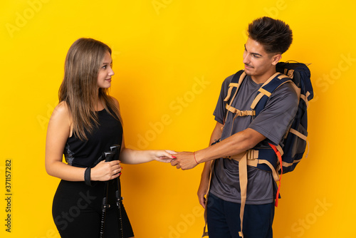 Young mountaineer couple with a big backpack isolated on yellow background handshaking after good deal