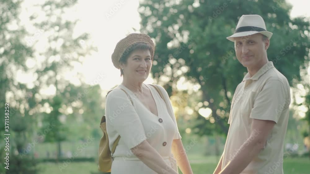 Loving mid-adult man and woman standing in sunrays holding hands and smiling at camera. Portrait of happy Caucasian couple posing in sunlight at sunset in summer park. Dating outdoors. Loving mid-adult man and woman standing in sunrays holding hands and smiling at camera. Portrait of happy Caucasian couple posing in sunlight at sunset in summer park. Dating outdoors.