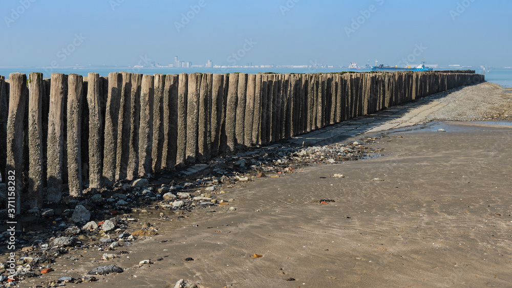 A breakwater made of two rows of wooden poles and large ships and buildings at the background.