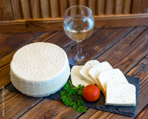 View of cheese head and cheese slicing on a black slate board served with tomatoes and fresh greens. Glass of dry white wine. Homemade cheese. Food concept. Selective focus. photo