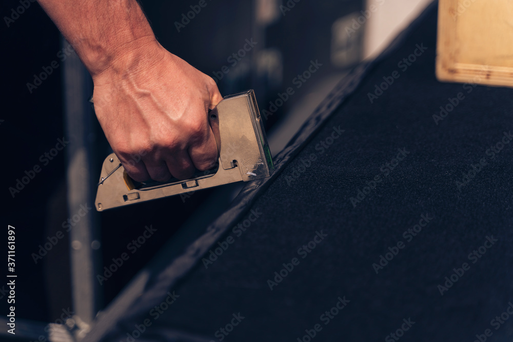 Upholstery workshop. Upholstery stapler working process. Restoring old sofa or stage podium upholstery. Man hands working. Man working with pneumatic stapler. - obrazy, fototapety, plakaty 