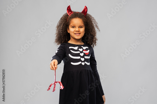 halloween, holiday and childhood concept - smiling african american girl in black costume dress with red devil's horns and trident over grey background