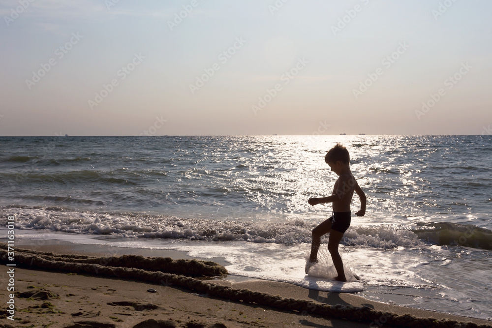 Little boy is playing on seashore in backlight at sunrise. Light toning.