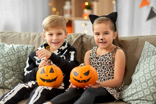 halloween, holiday and childhood concept - smiling little boy and girl in costumes with jack-o-lantern pumpkins having fun at home