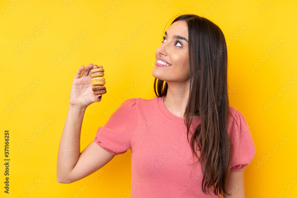 Young brunette woman over isolated yellow background holding colorful French macarons and looking up while smiling Young brunette woman over isolated yellow background holding colorful French macarons and looking up while smiling