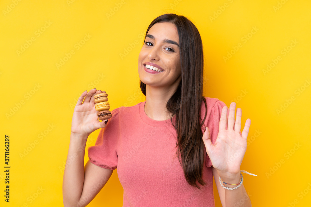 Young brunette woman over isolated yellow background holding colorful French macarons and saluting Young brunette woman over isolated yellow background holding colorful French macarons and saluting