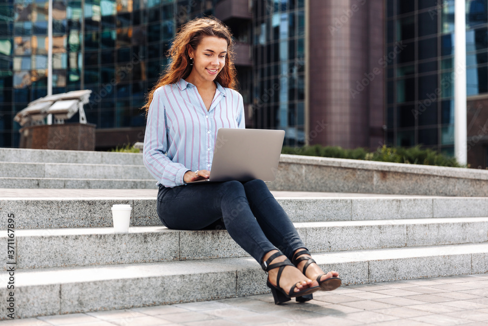 Smiling businesswoman working at laptop outdoors in front of business center sitting on steps