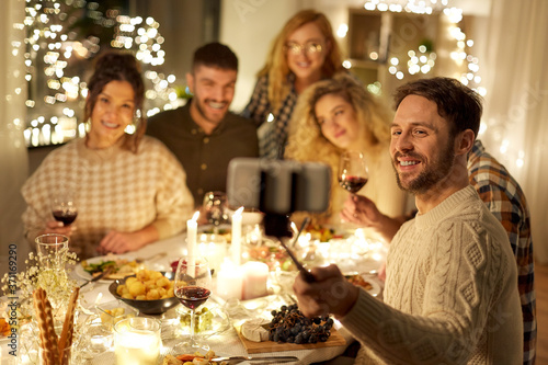 holidays  celebration and people concept - happy friends drinking non-alcoholic red wine and taking picture with smartphone on selfie stick at home christmas dinner party