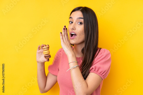Young brunette woman over isolated yellow background holding colorful French macarons and whispering something