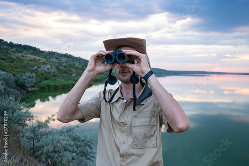 Young male traveler or scientist with binocular and photo camera outdoors near river. 