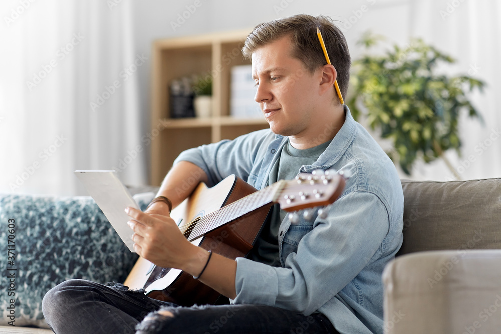 leisure and people concept - young man or musician with guitar and music book sitting on sofa at home