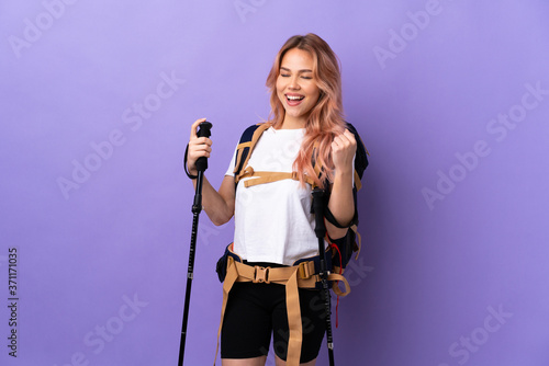 Teenager girl with backpack and trekking poles over isolated purple background celebrating a victory