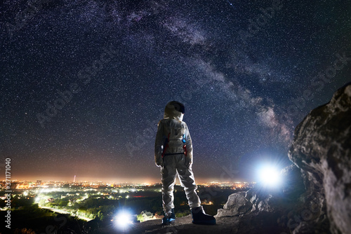 Mission specialist astronaut standing on rocky hill and looking at beautiful starry sky with milky way. Space traveler wearing white space suit and helmet. Concept of human space exploration.