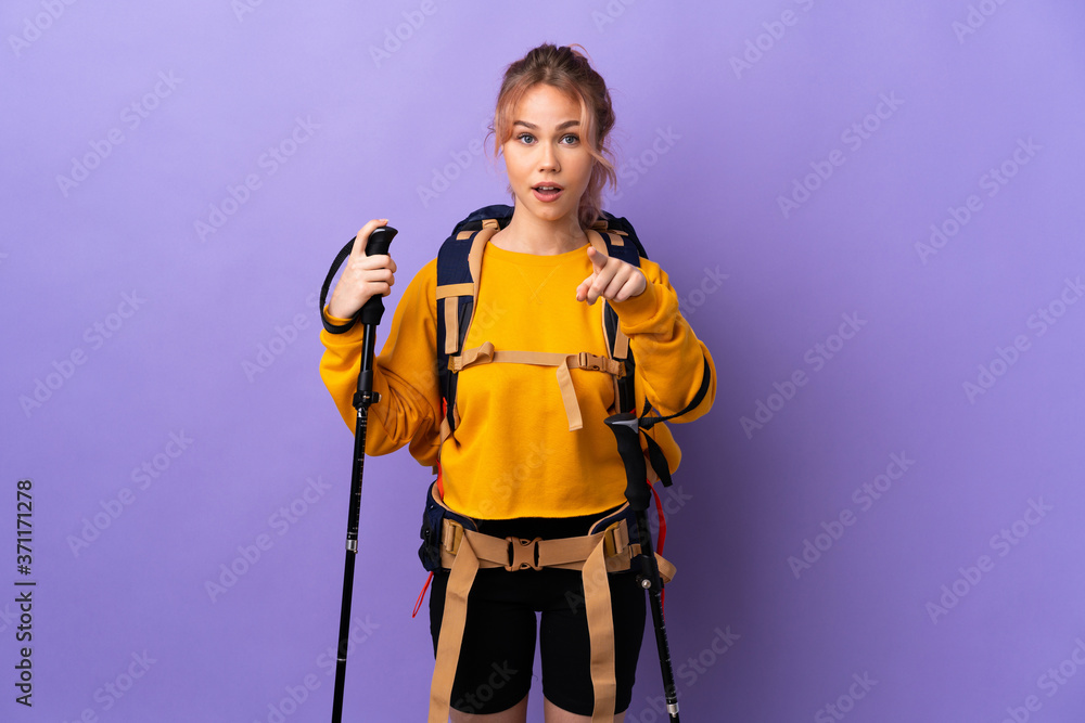 Teenager girl with backpack and trekking poles over isolated purple background surprised and pointing front