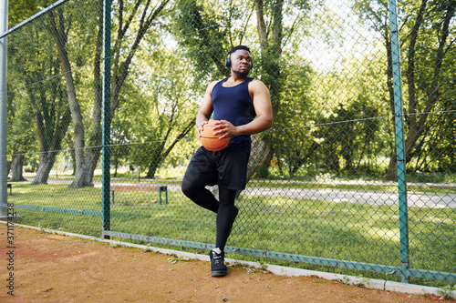 African american man with wireless headphones takes a break and leaning on the metal mesh with ball on the court outdoors