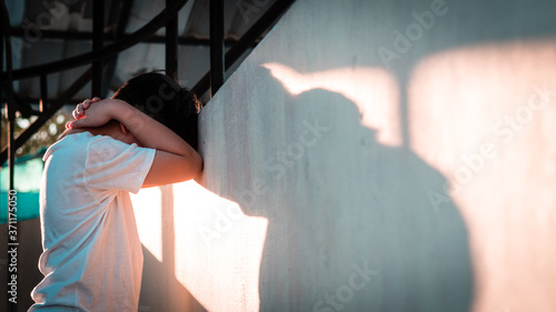 Depressed, frustrated and overwhelmed Asian teenage boy stand alone and lean his head against wall with casting shadow, feeling worthless, stressed, guilty, hopeless and having suicidal thought. photo