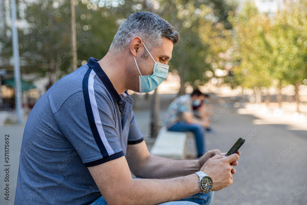 Obraz premium Four men with medical masks watching their cellphones sitting on benches far apart in the street. Social distancing concept.