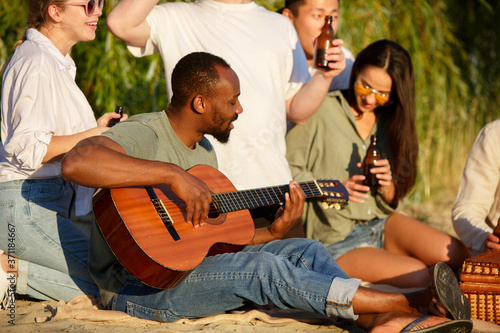 Guitar playing. Group of friends clinking beer glasses during picnic at the beach in sunshine. Lifestyle, friendship, having fun, weekend and resting concept. Looks cheerful, happy, celebrating © master1305