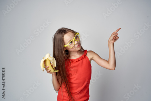 little girl in glasses and a red dress with a banana in her hands light background