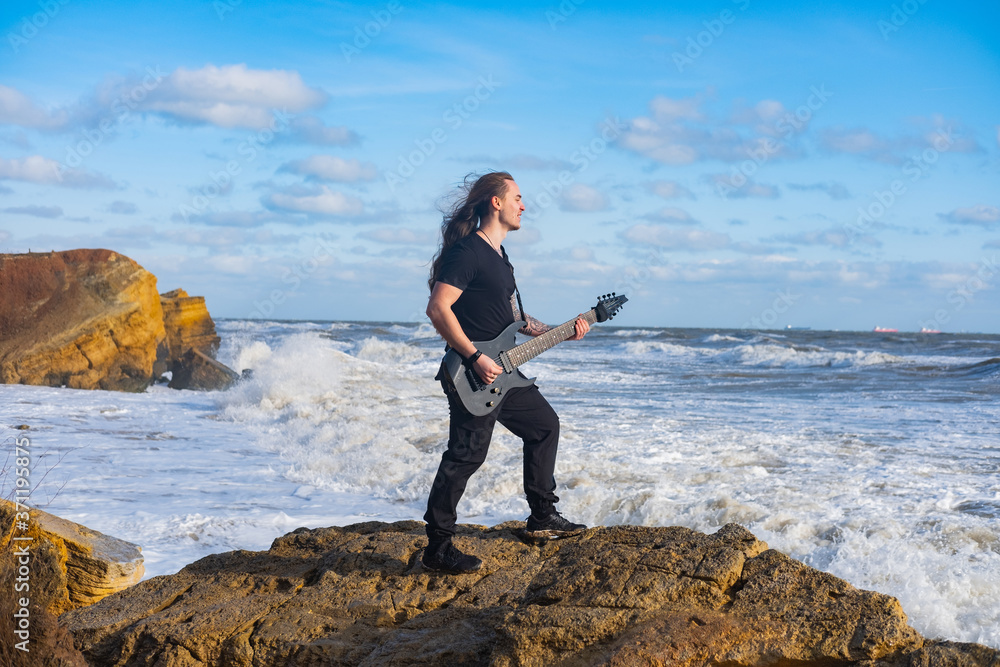 Young male in black and long hair play on guitar on the beach