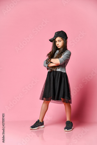 Tableau sur toile Portrait of a little hipster girl in a dark dress with a baseball cap and sneake