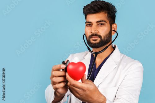 Indian / Asian doctor with a stethoscope and a red heart in his hands, in a white coat on a blue background. cardiologist © Nana_studio