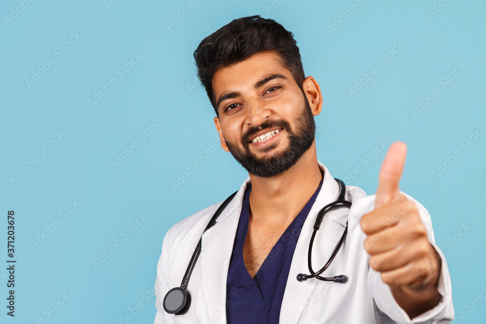 young Indian / Asian doctor with stethoscope in white coat showing thumb up on blue background. young Indian / Asian doctor with stethoscope in white coat showing thumb up on blue background.