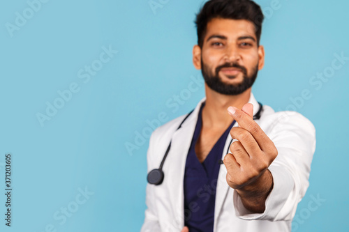 handsome young Indian / Asian doctor with stethoscope in white coat shows heart sign with fingers on blue