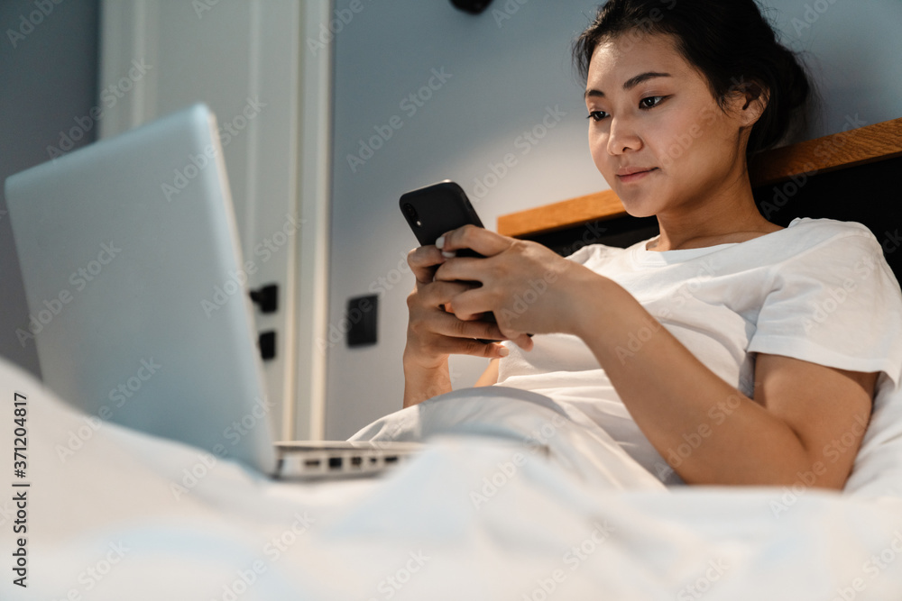 Smiling beautiful young asian woman working on a laptop while sitting in bed, using mobile phone Smiling beautiful young asian woman working on a laptop while sitting in bed, using mobile phone