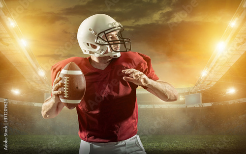 American football sportsman player with ball in action on stadium under lights of background. Sport, proud footballer in white helmet and red t-shirt ready to play.  © Andrii IURLOV