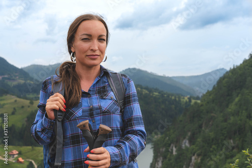 Portrait of Young mountaineer woman with a big backpack, climbing sticks and blue shirt. Mountain lake in backgorund.