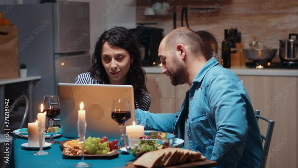 Excited caucasian lover couple dining together and using laptop. Adults sitting at the table, searching, browsing, surfing, using technology, enjoying the meal in the dining room Excited caucasian lover couple dining together and using laptop. Adults sitting at the table, searching, browsing, surfing, using technology, enjoying the meal in the dining room