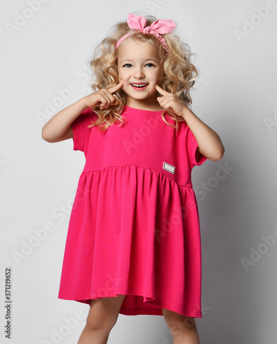 Cute smiling blonde kid girl with curly hair in stylish girlish pink dress and headband holds fingers at her cheeks