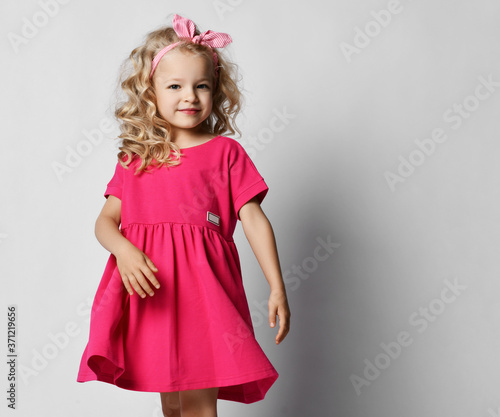 Little blonde kid girl with curly hair in stylish girlish pink dress and headband with bow is dancing spinning