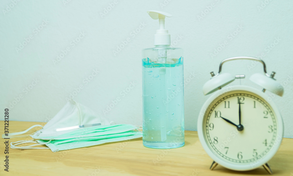 The sanitizer with a medical mask and a white clock is placed on a wooden floor in a light blue wall background. Surveillance of COVID-19 outbreaks with medical masks, including hand washing gel. The sanitizer with a medical mask and a white clock is placed on a wooden floor in a light blue wall background. Surveillance of COVID-19 outbreaks with medical masks, including hand washing gel.