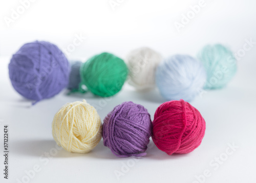 Three small balls of yarn in a row on a light surface, with other colored balls of yarn in the background. Knitting or crochet materials up close with side light.