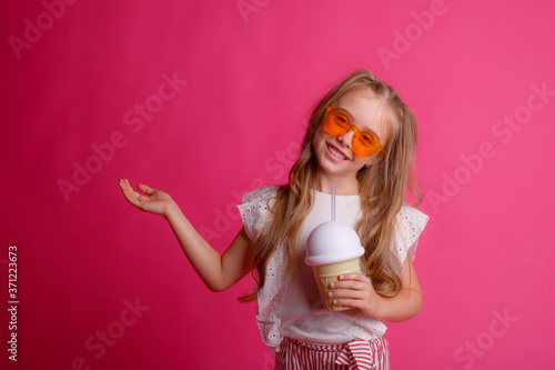 portrait of a little blonde girl in sunglasses on pink with a glass jar of cooling cocktail.