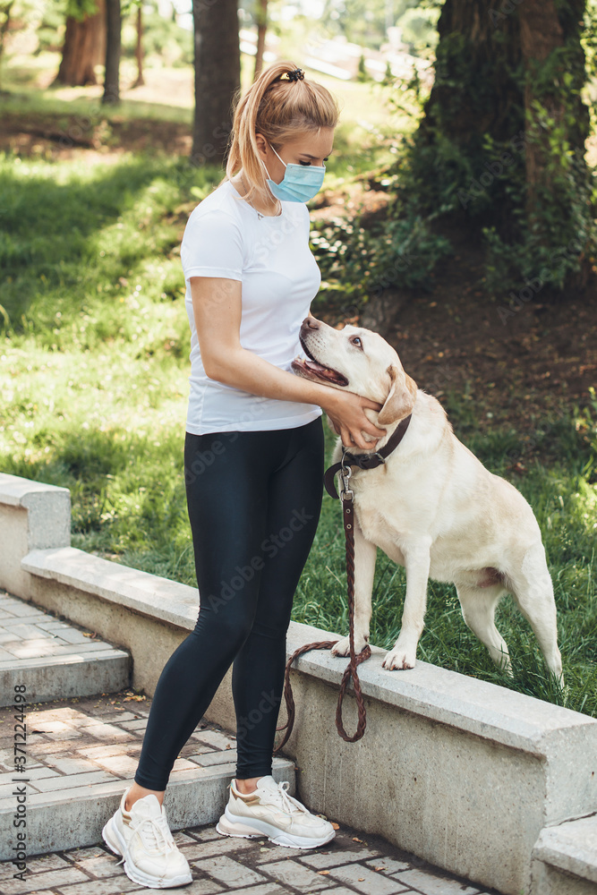 Caucasian blonde lady with medical mask on face walking with her dog outside in the park